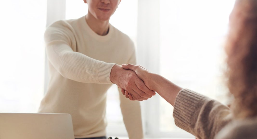 A person sitting at a desk, shaking the hand of a prospective employer, as they are interviewing for a high-paying job that pay $75,000 or more per year