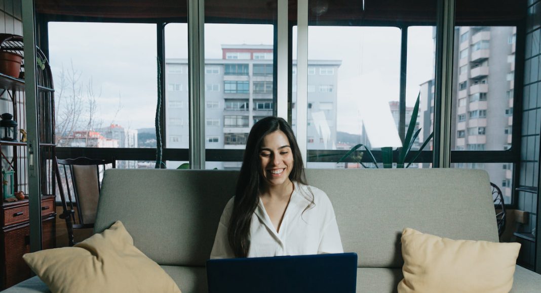 Woman sits in the middle of there couch with orange pillows on each side and looks at a laptop that is on their lap.