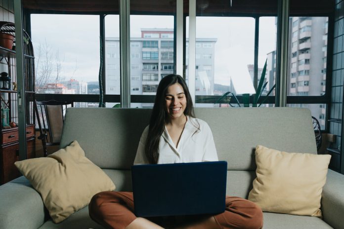 Woman sits in the middle of there couch with orange pillows on each side and looks at a laptop that is on their lap.